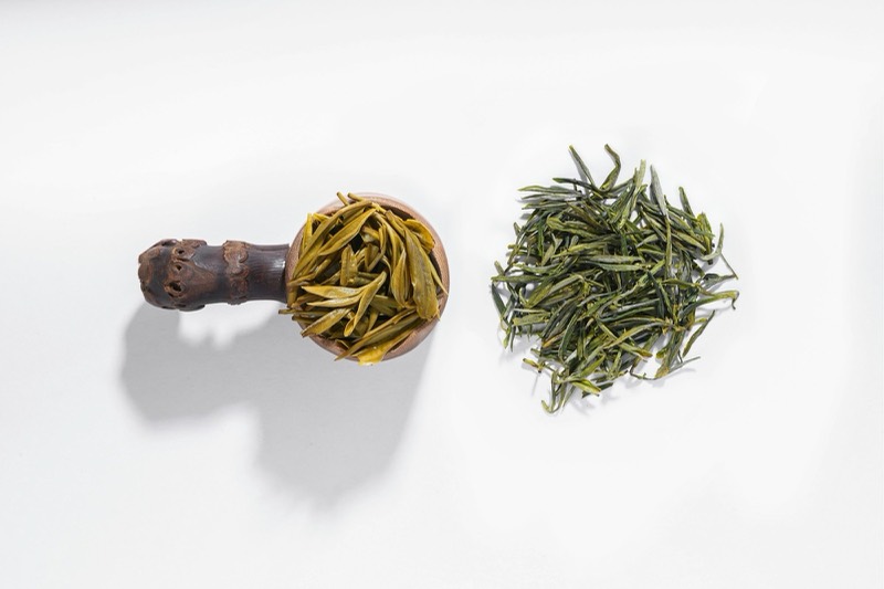 Dried green tea leaves in a wooden scoop beside a small pile, on white